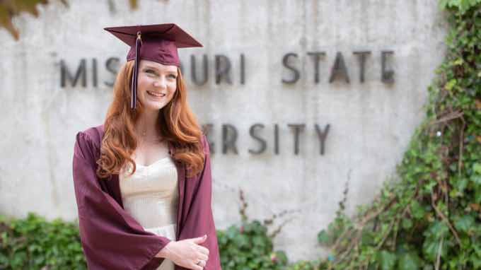 Student in cap and gown standing in front of ivy-covered wall