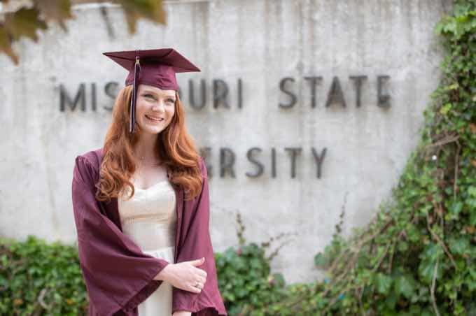 Student in cap and gown standing in front of ivy-covered wall